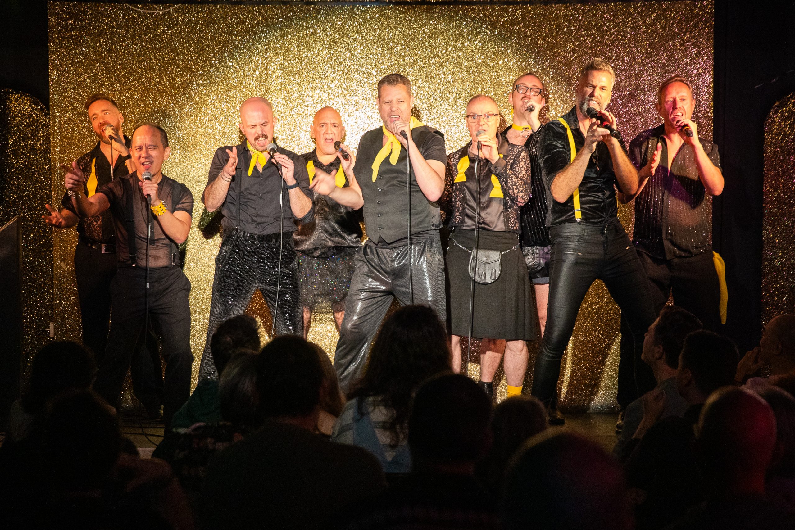 A group of 9 singers dressed in black with yellow accents performing on a stage with a gold glittery background and a spotlight on them.
